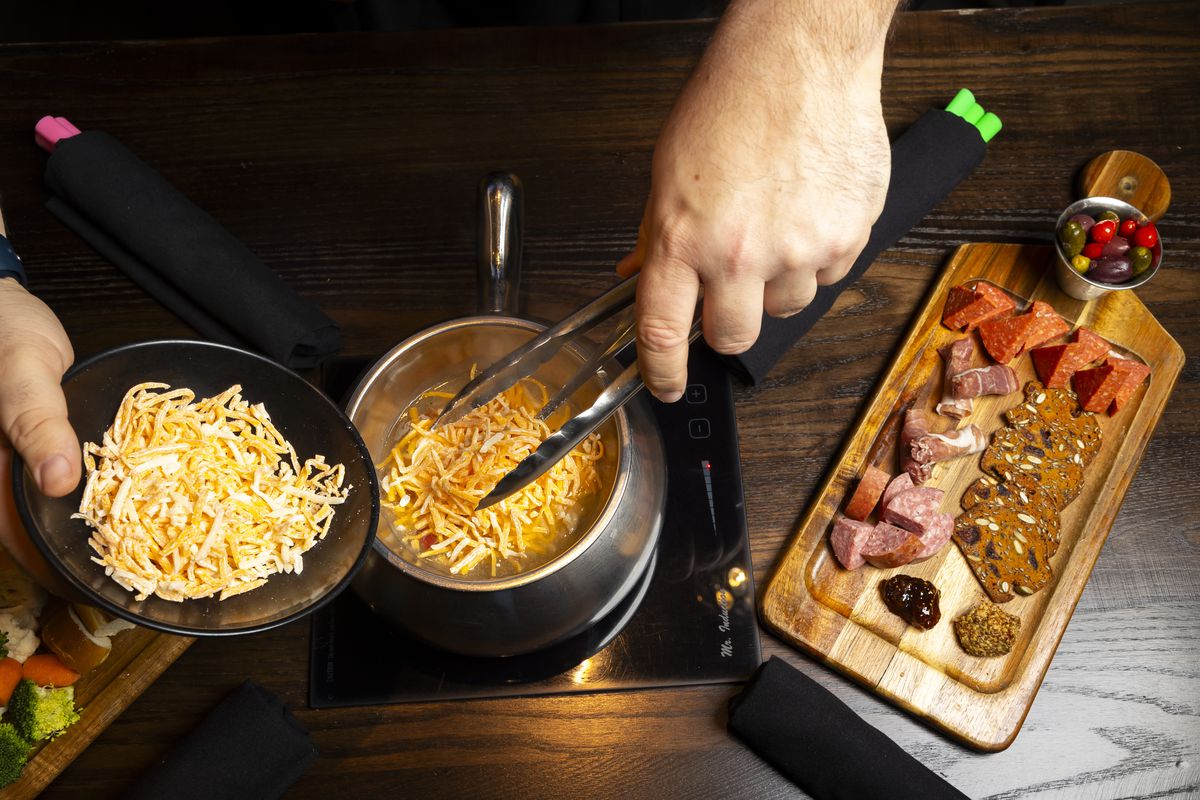 An overhead shot of a person using tongs to add shredded cheese to a fondue pot. Alongside the pot a wooden serving board holds a cup of olives, chunks of meat, and crackers.