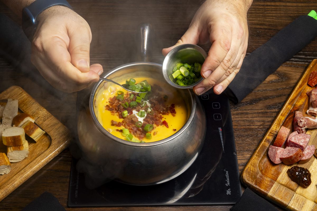 A person spoons scallions into a fondue pot filled with cheese and topped with bacon bits and sour cream. 