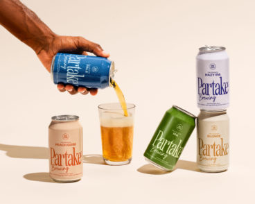 A hand pours a can of Partake non-alcoholic beer into a glass that is surrounded by other cans of Partake