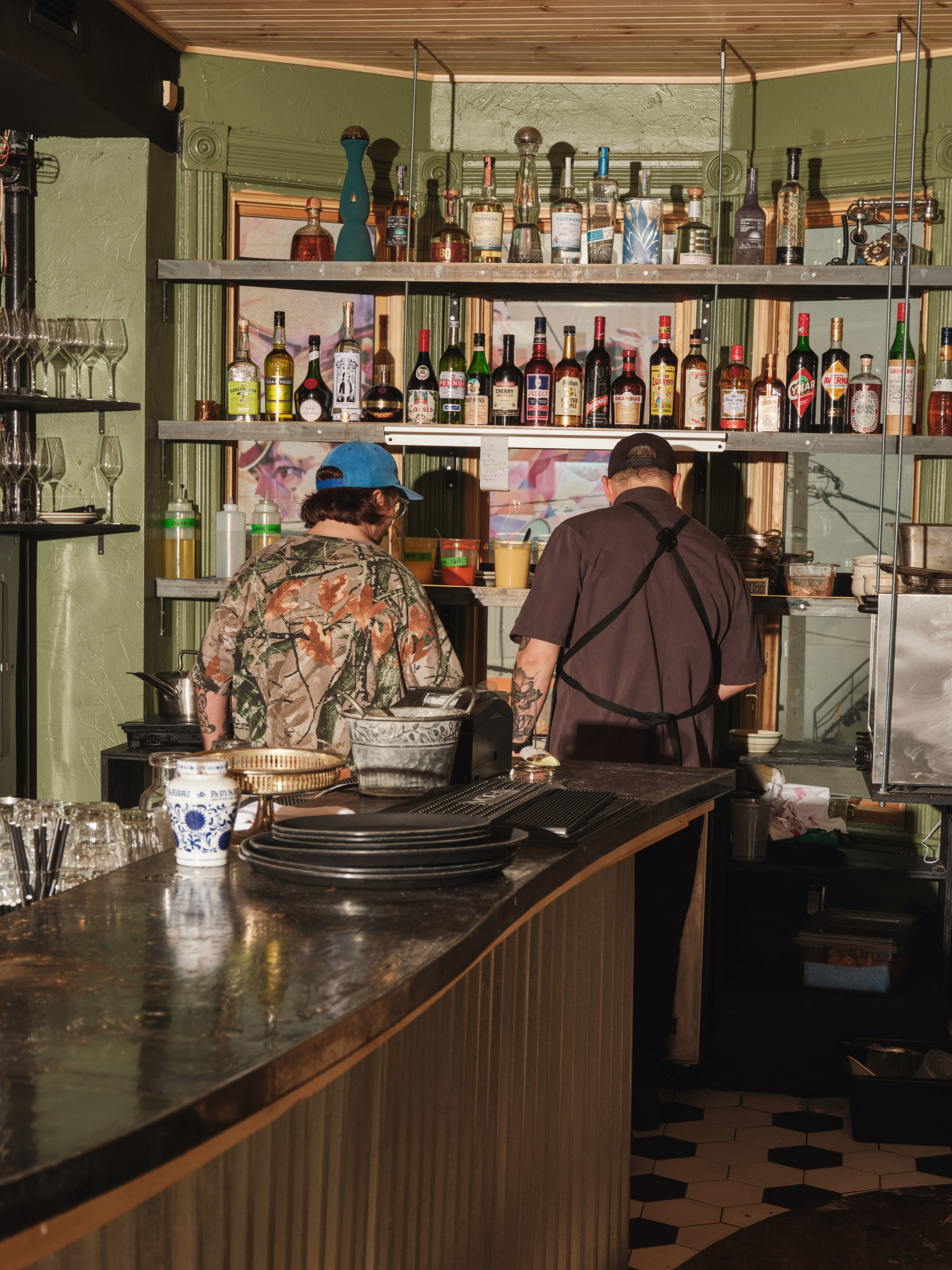 Two men, both wearing hats and one wearing an apron, work behind a bar and under two shelves lined with liquor bottles