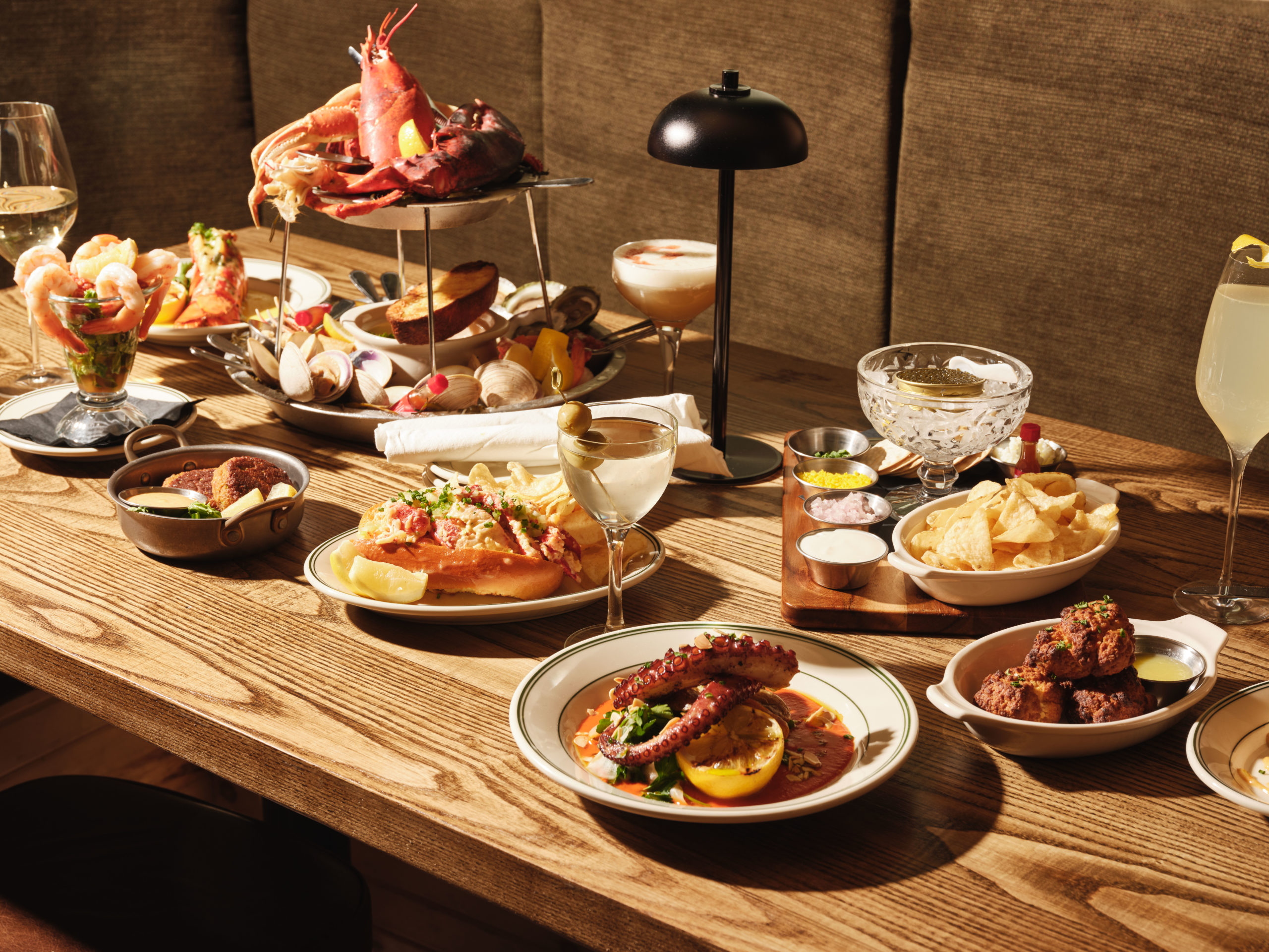 A table is spread with all kinds of seafood dishes, including a lobster-topped tower, and cocktails